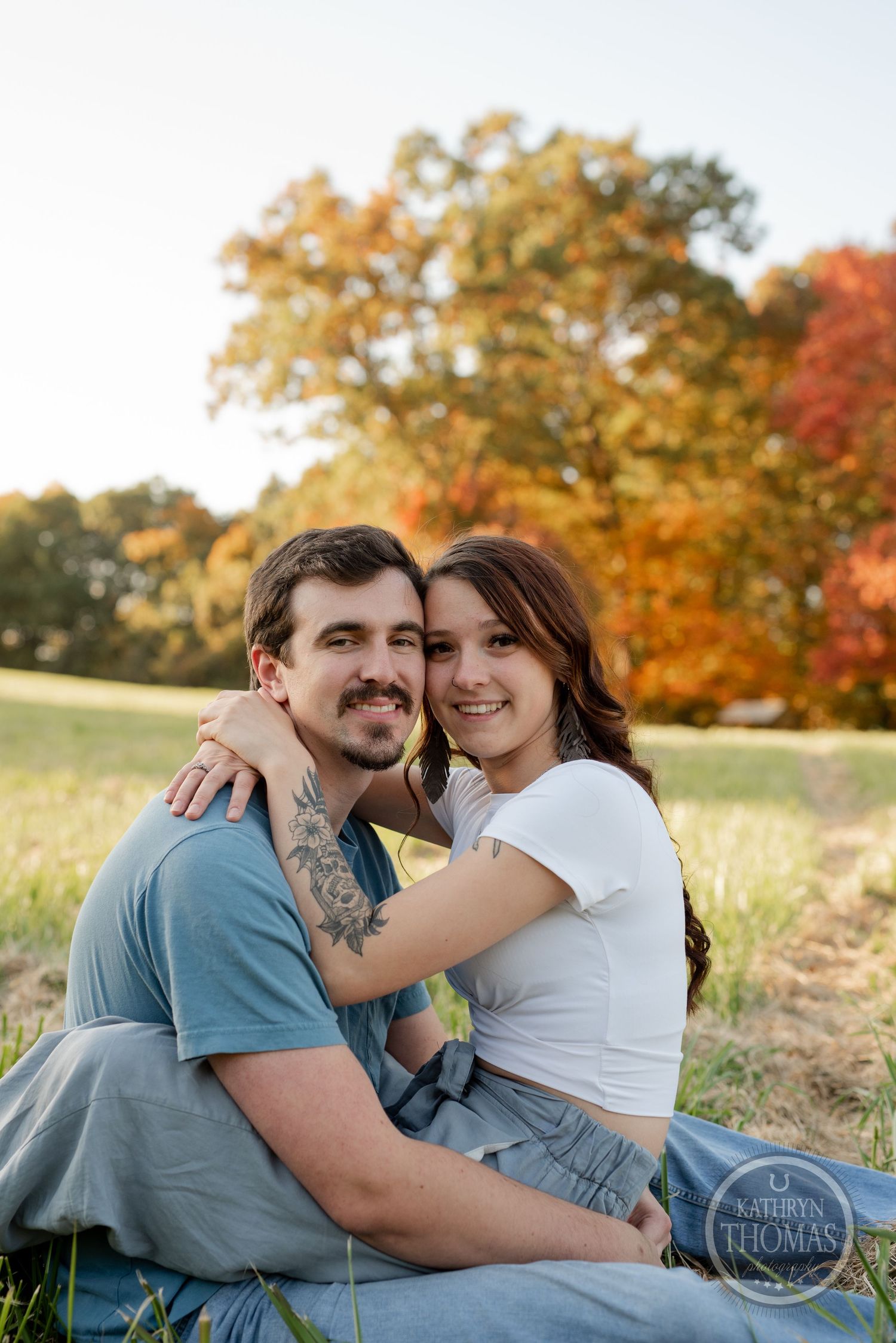 A couple shares a tender embrace while sitting in a field with fall colors in the background.