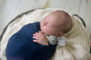 Sleeping newborn swaddled in navy blue wrap with small plush toy on cream blanket.