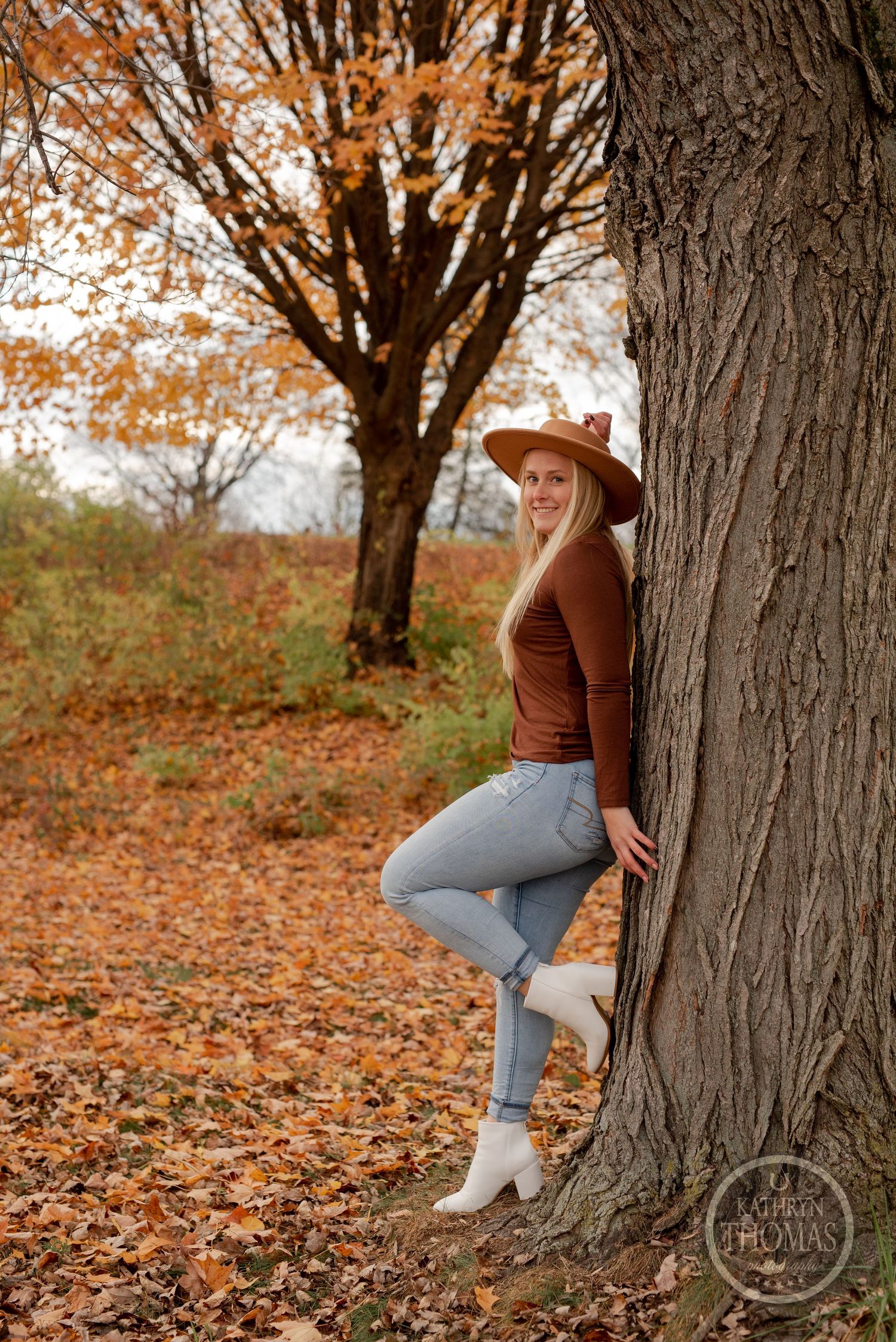 Person in brown sweater and jeans leans against large tree trunk surrounded by fall foliage.