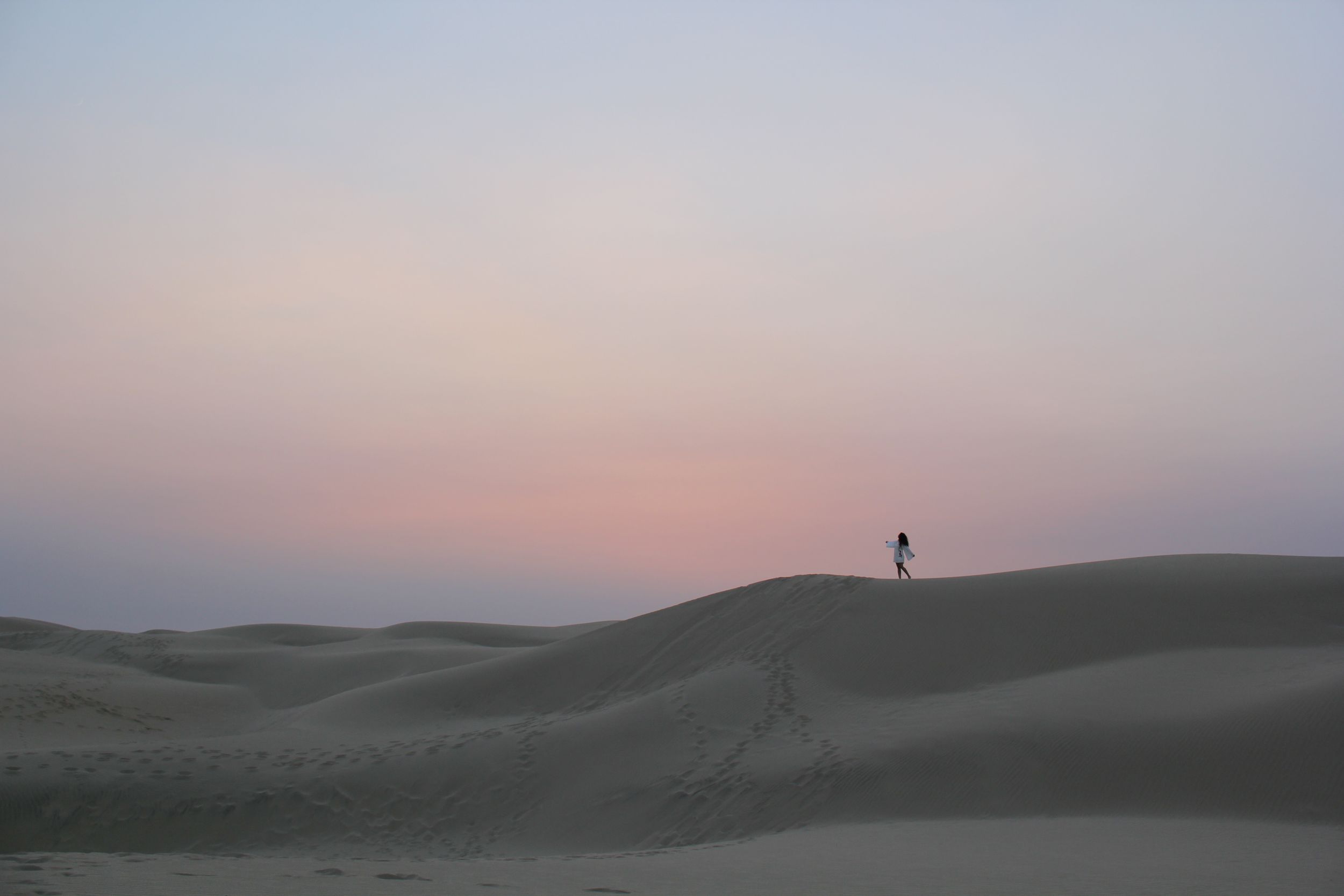 Lone figure stands on sand dune against pastel sunset sky in CA.