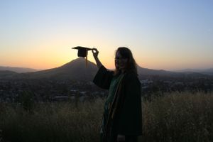 A silhouette holds up a graduation cap against a sunset mountain backdrop.