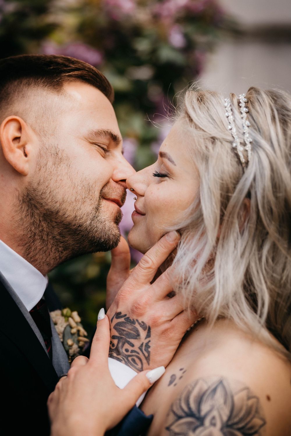 Close-up wedding photo of a couple sharing an intimate moment with visible tattoos and floral backdrop.