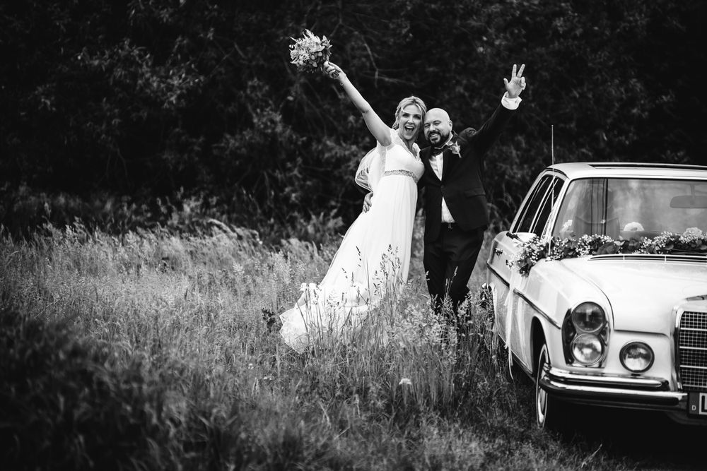 Black and white wedding photo of celebrating newlyweds next to vintage car in field.