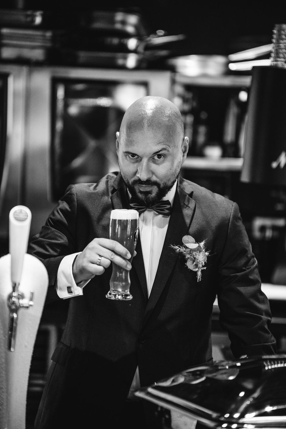 Groom in black suit sips from a glass at a bar counter in a dimly lit setting.