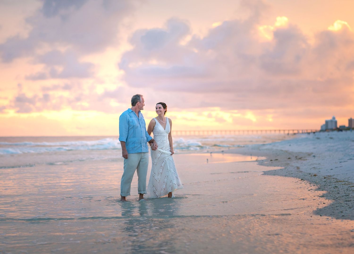 Wedding couple walking on beach during golden hour with pier in background.