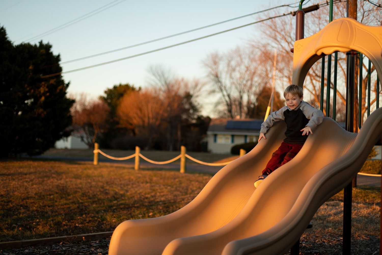 Child gliding down orange-lit playground slide during sunset.