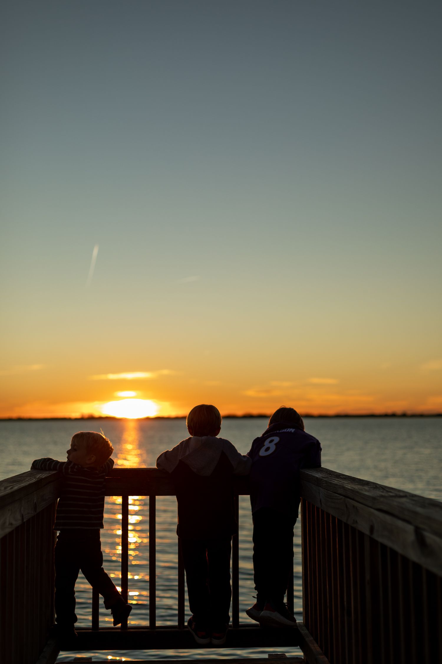Three silhouettes stand on pier watching sunset over calm water.
