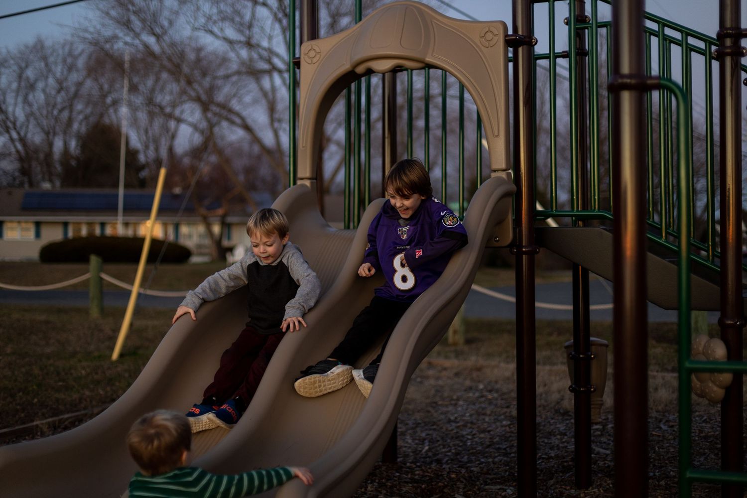 Two children sliding down playground equipment in evening light.