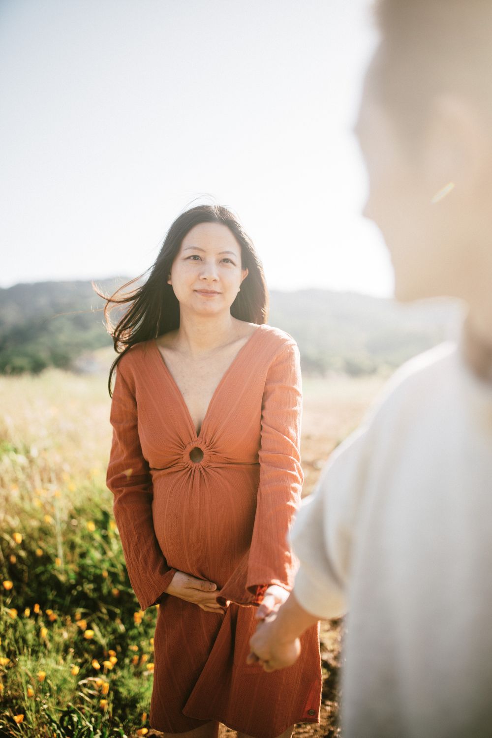 An expectant mother in a rust-colored dress stands in a sunlit field.