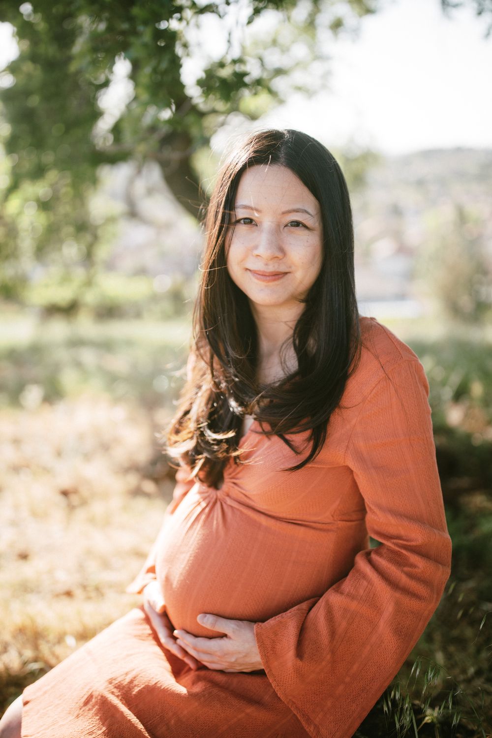 A glowing maternity portrait of a woman in a rust-colored dress amid nature.