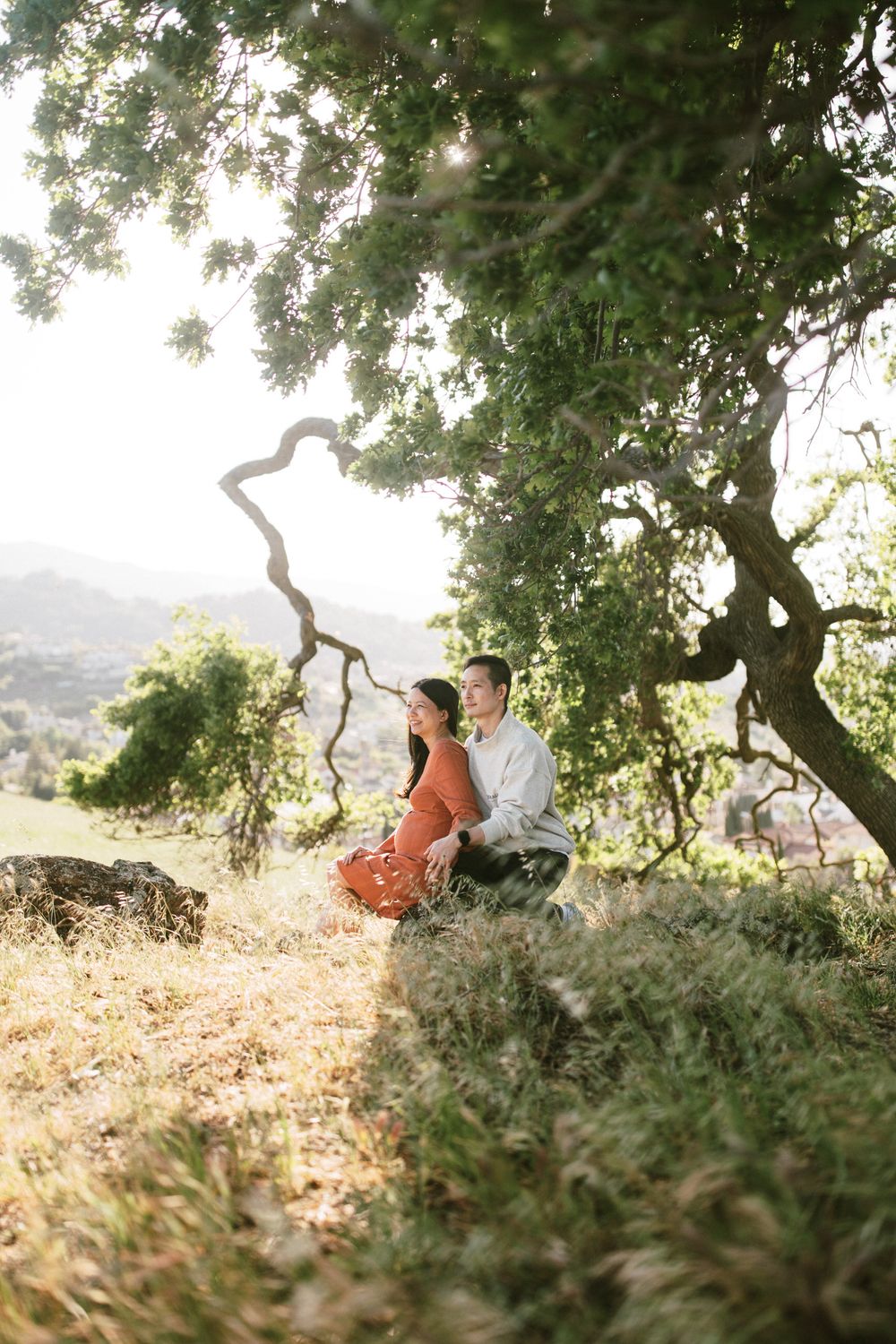 A couple sits under a twisted tree branch at sunset in a serene hillside setting.