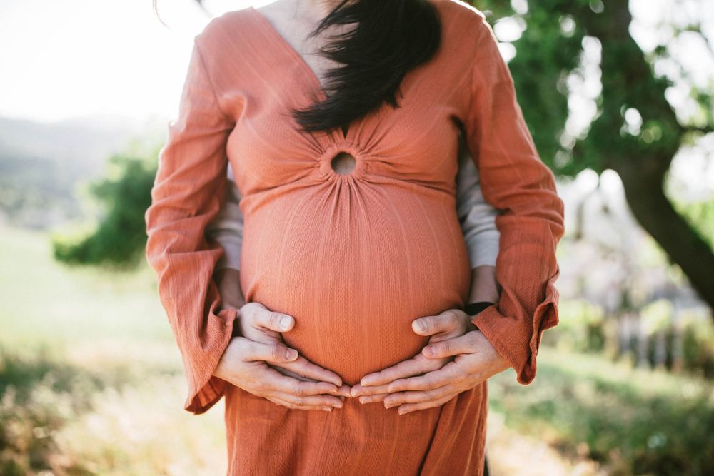 Close-up of hands cradling a pregnant belly in a rust-colored dress outdoors.
