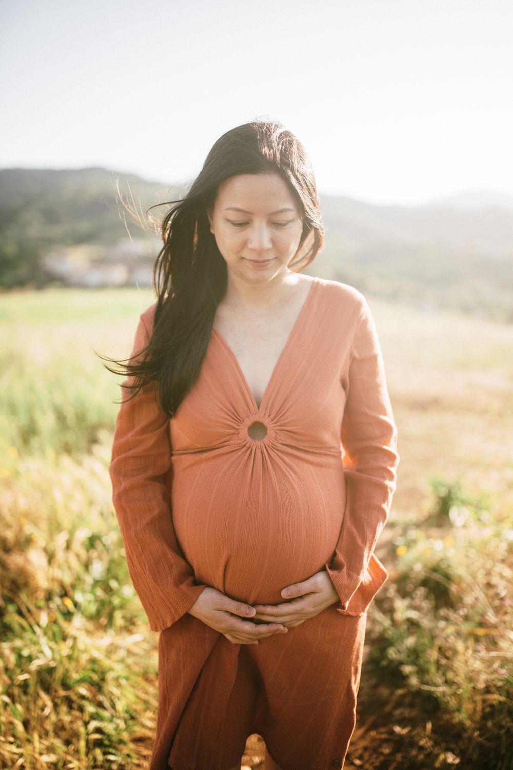 Expectant mother in rust-colored dress poses in golden sunset light with rolling hills.