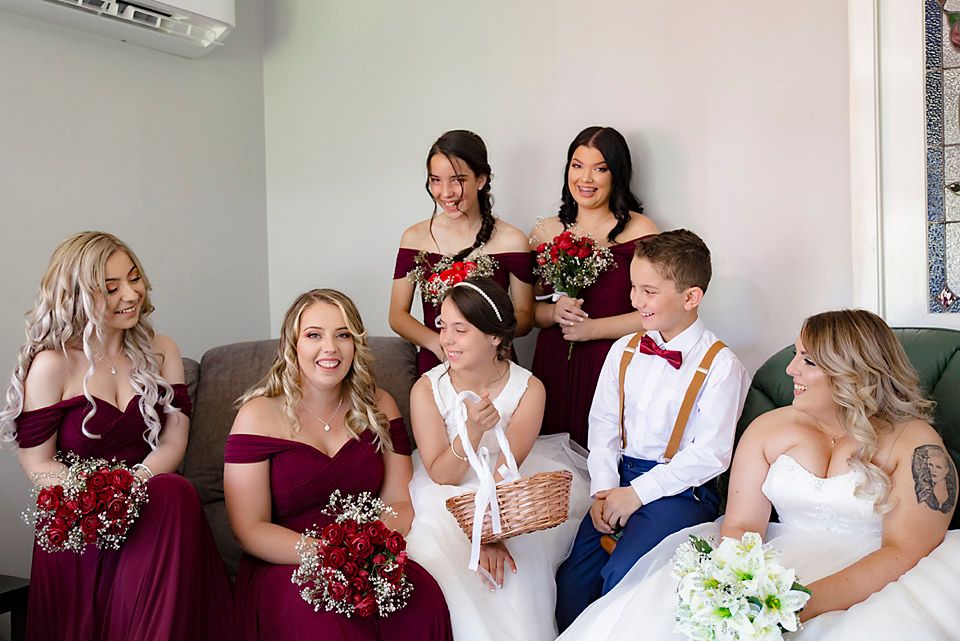 Bridal party getting ready in a bright room wearing burgundy dresses and white gown.