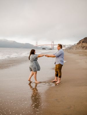 A couple dances playfully on the beach with the iconic Golden Gate Bridge visible in the background.