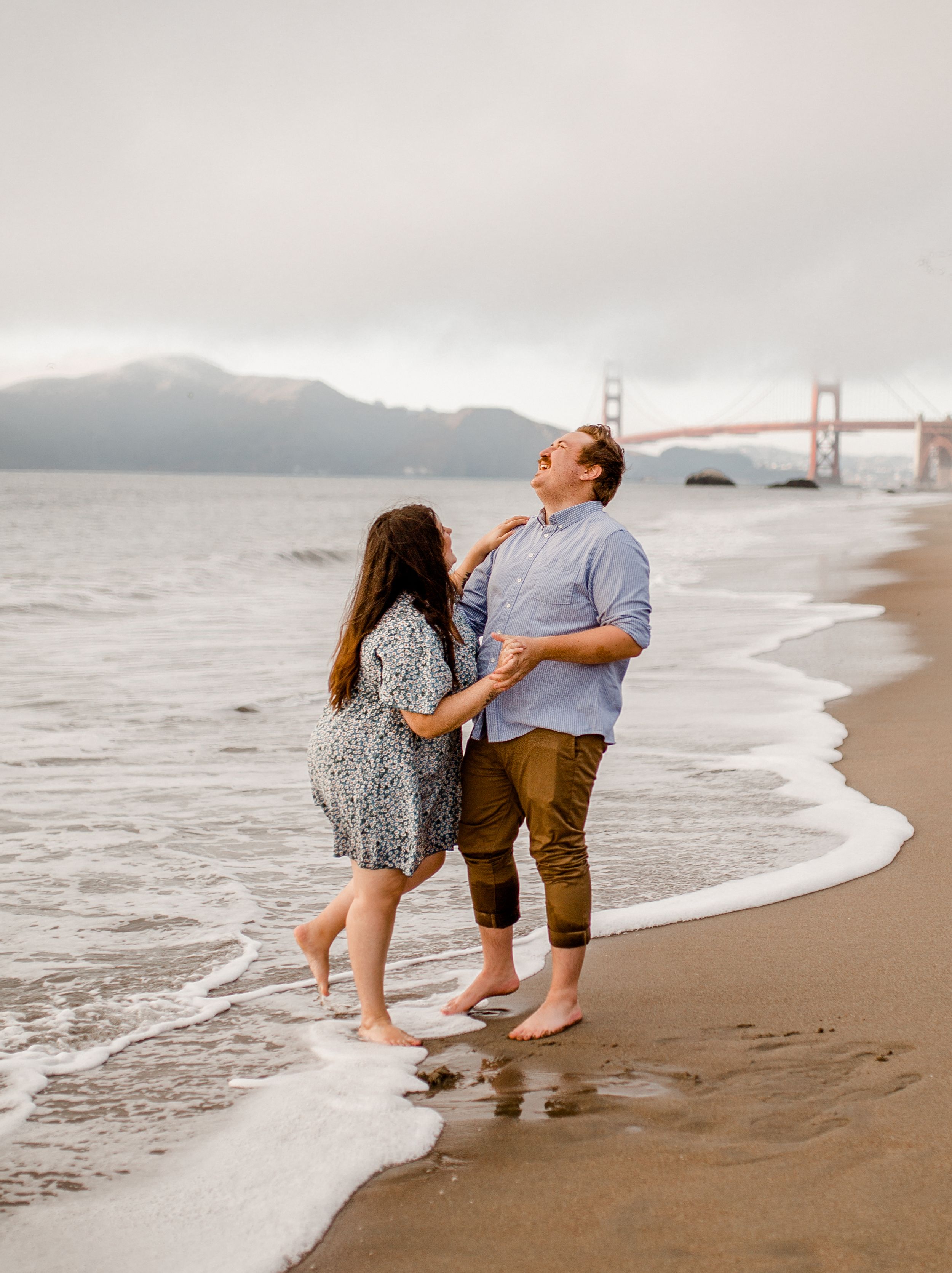A couple shares a joyful moment at the beach with San Francisco's Golden Gate Bridge in the distance.