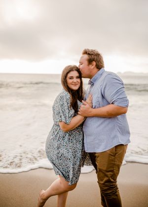 Romantic couple embrace on cloudy beach with waves in background.