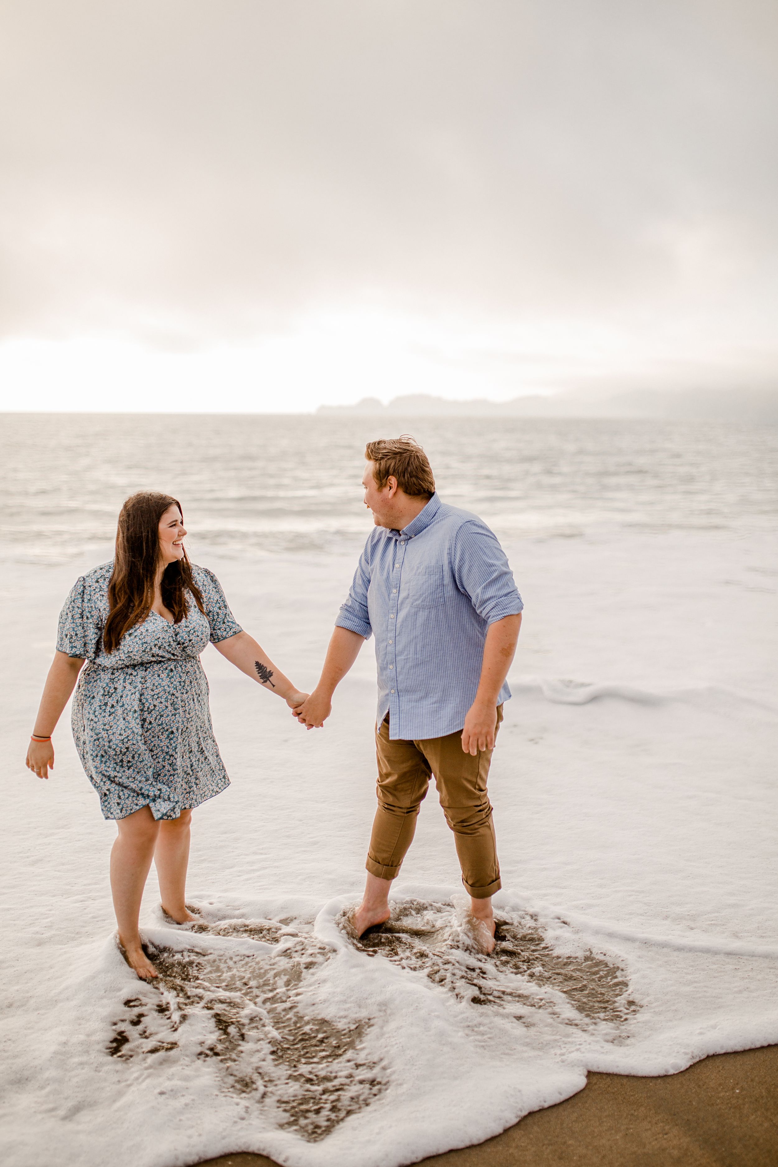 Couple holding hands while standing in ocean waves at sunset.