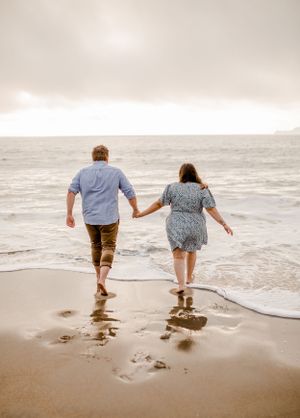 A couple holds hands while walking barefoot along the shoreline during a misty beach sunset.