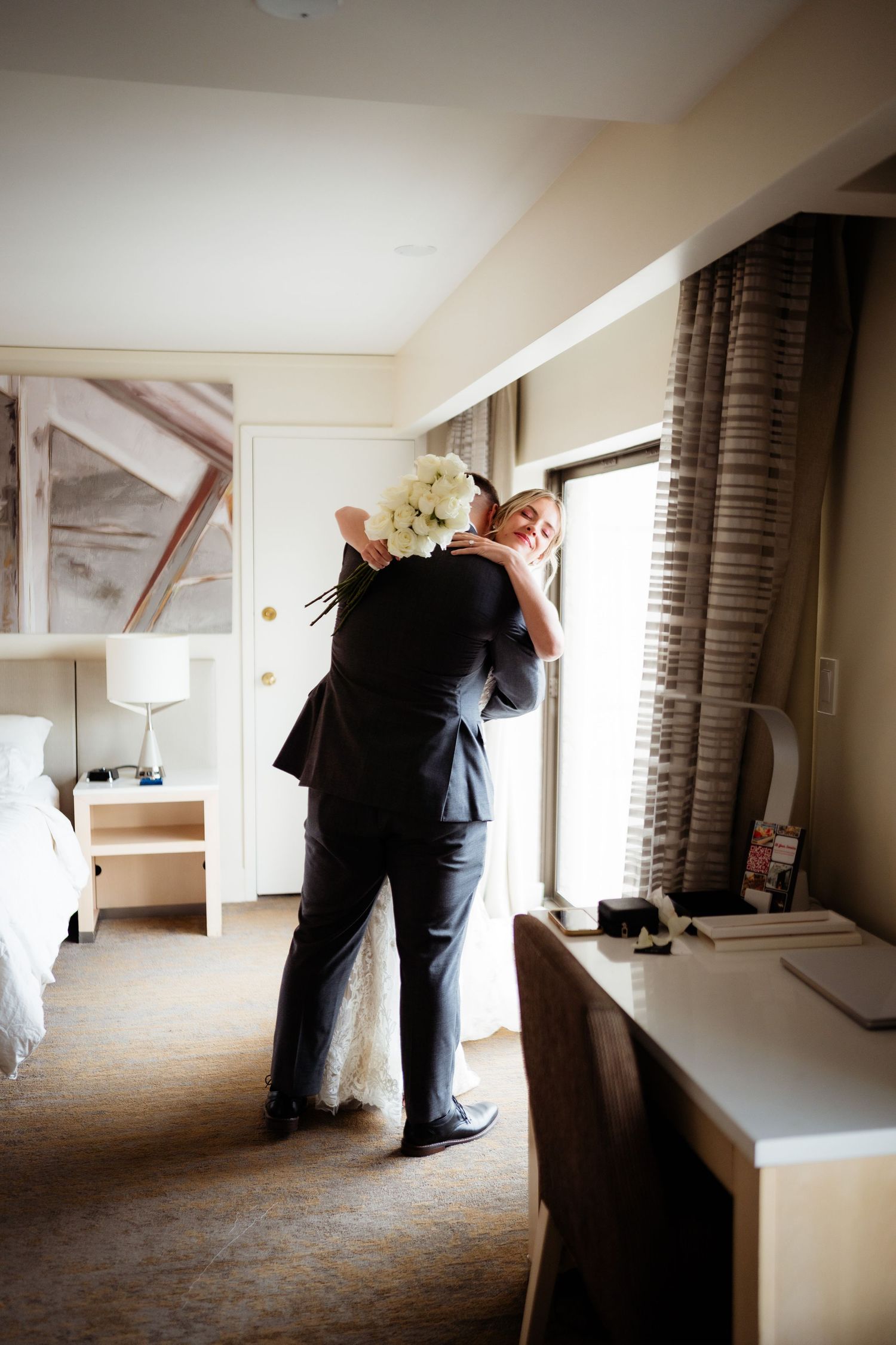 A couple embraces in a sunlit hotel room while wearing formal attire.
