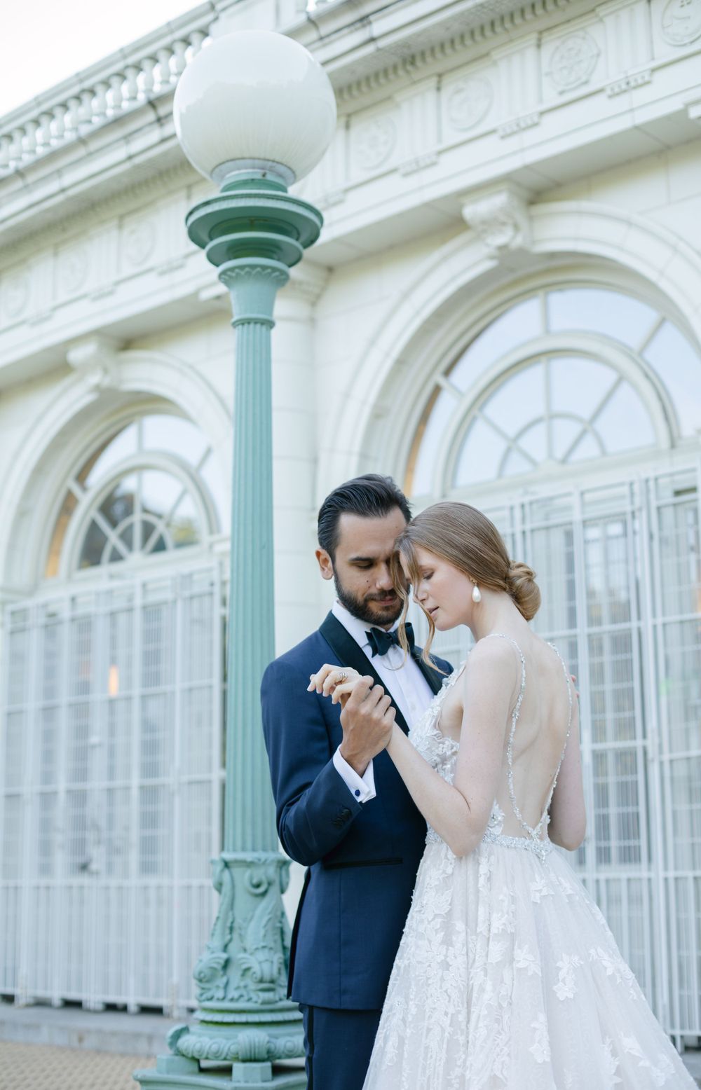A romantic wedding couple poses by an ornate white building with vintage lampposts.