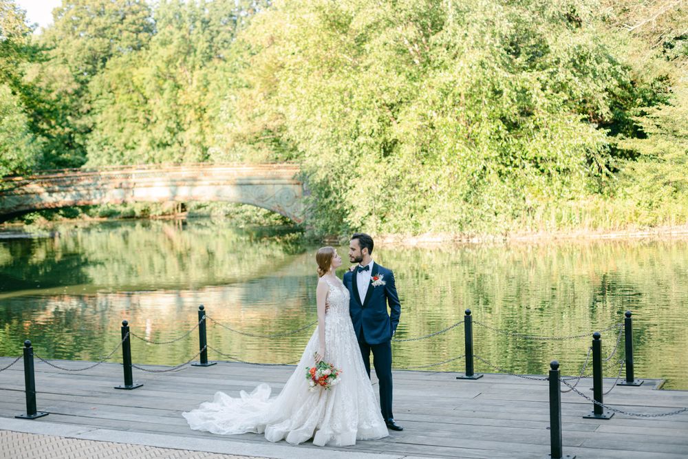Wedding couple standing on wooden dock overlooking serene lake surrounded by lush greenery.