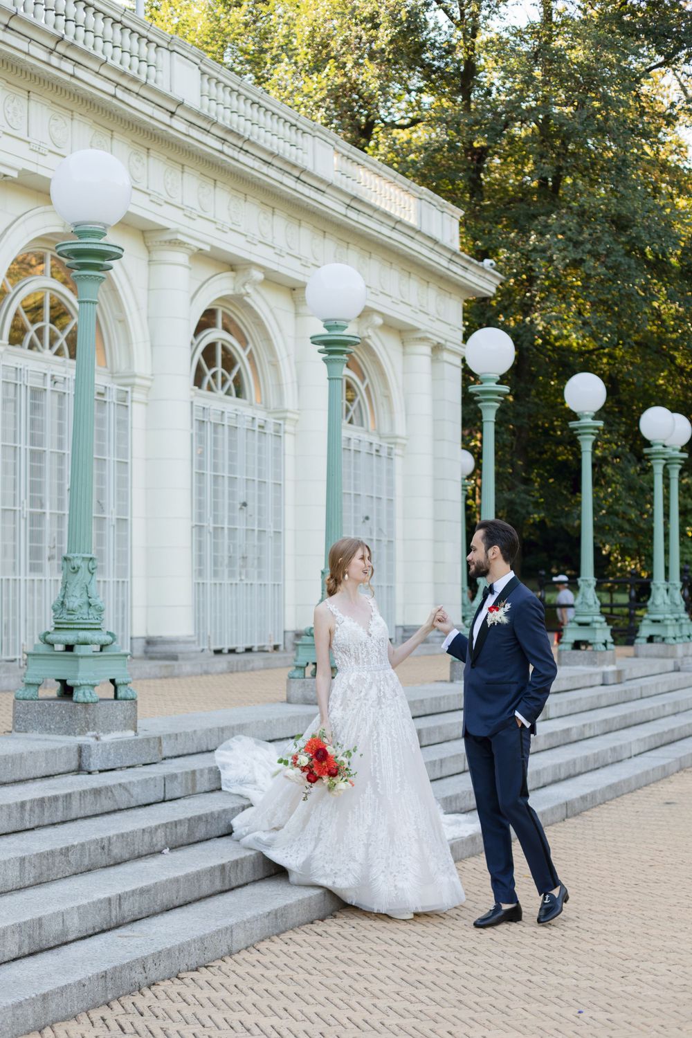 A wedding couple poses on stone steps of a grand white building with elegant lamp posts.