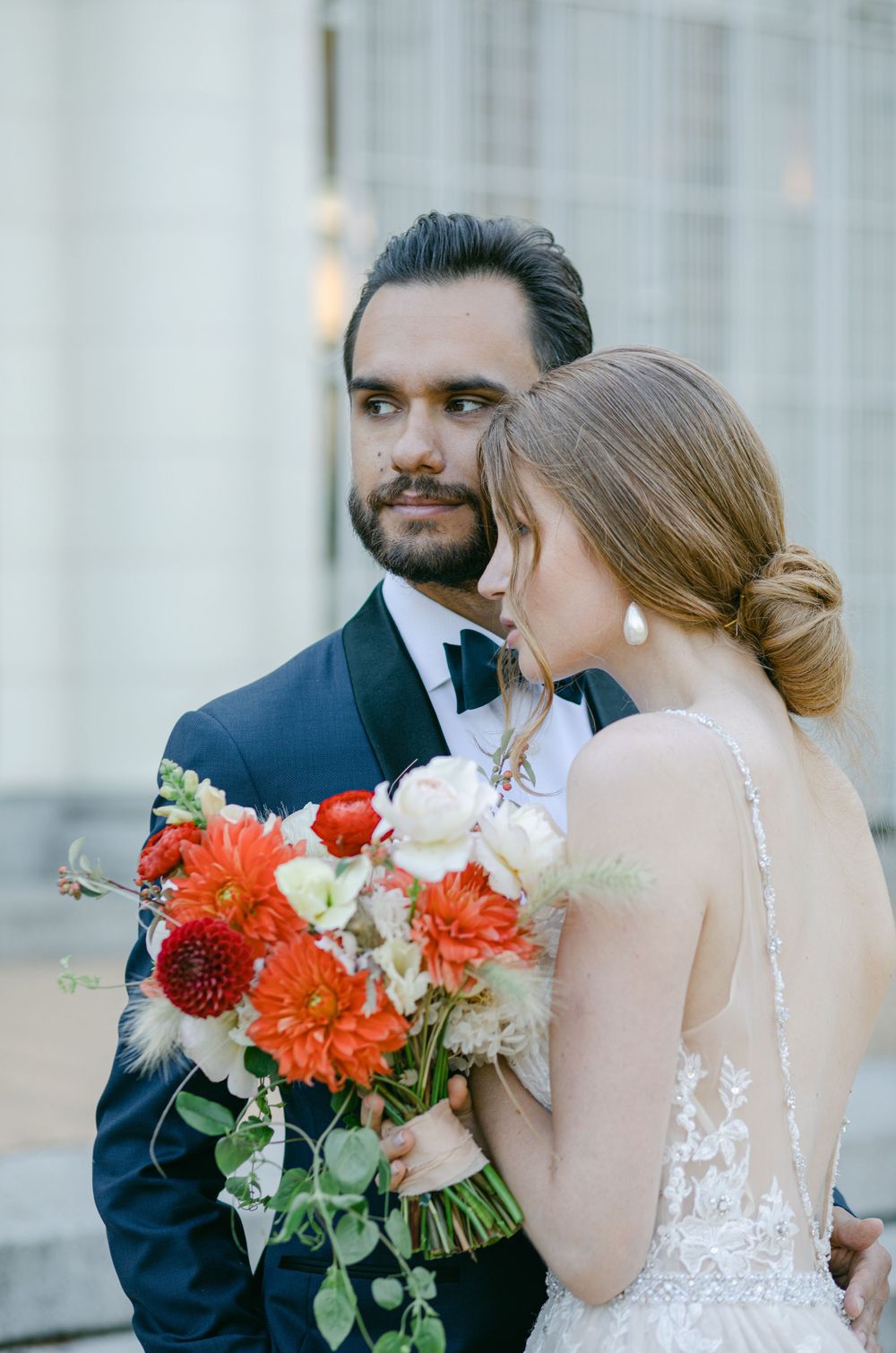 Romantic wedding portrait with vibrant orange and red bouquet against classic architectural backdrop.