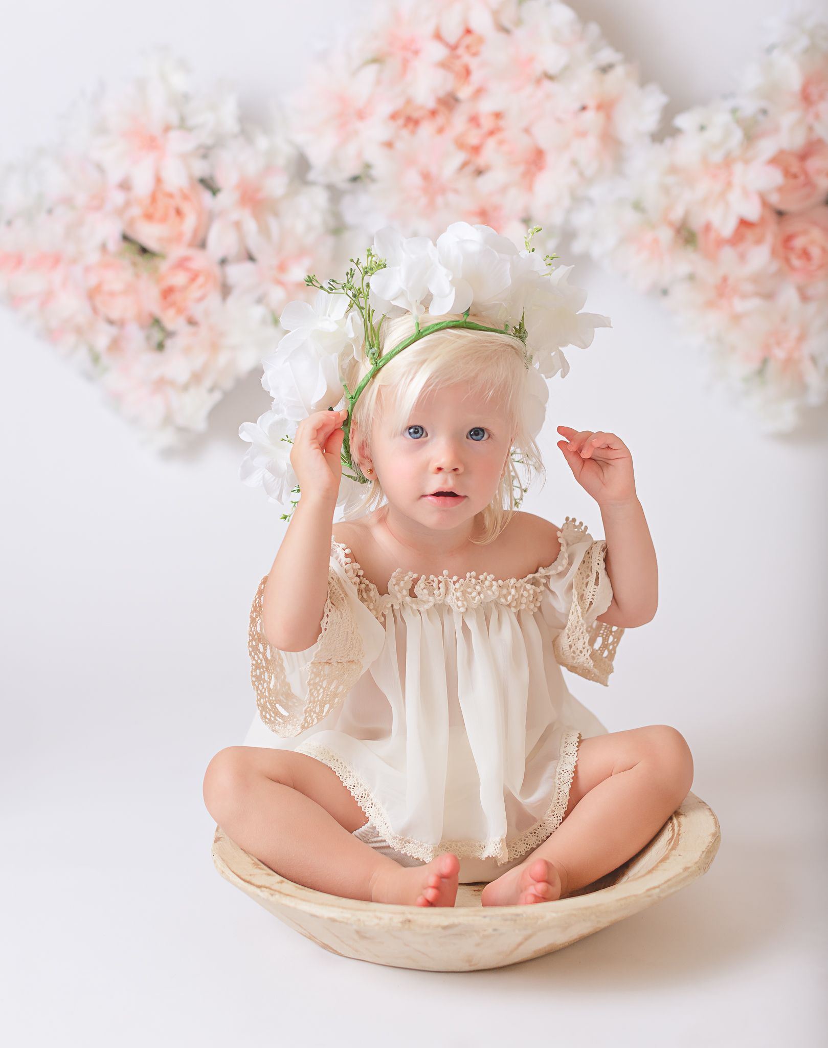 Baby in vintage cream dress posing with pink and white flowers in background.