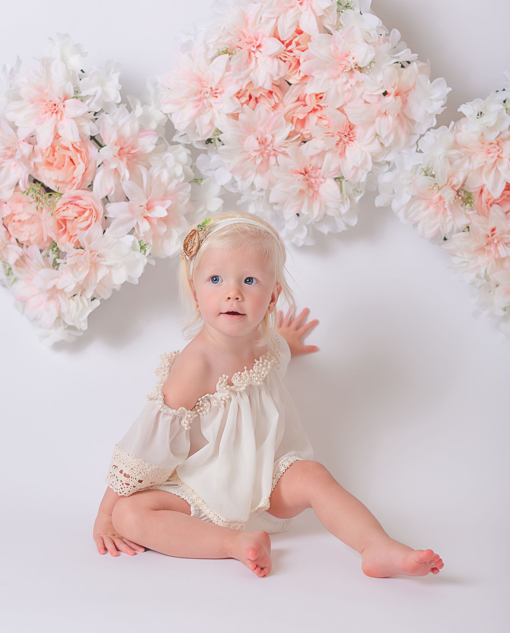 Baby in cream dress crawling under pink and white floral garland.