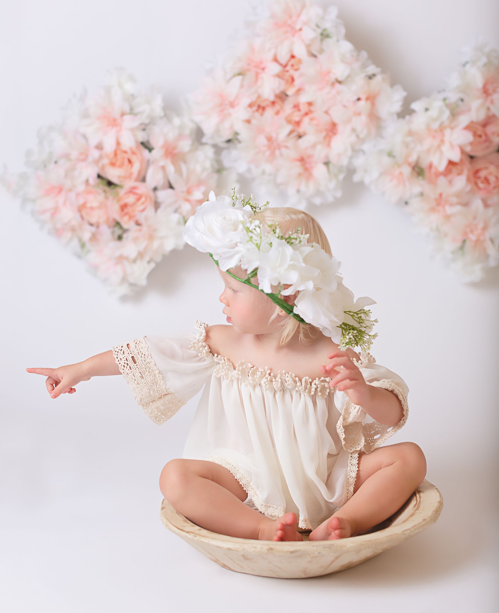 Baby in cream dress lying on white surface with delicate pink and white floral decorations.