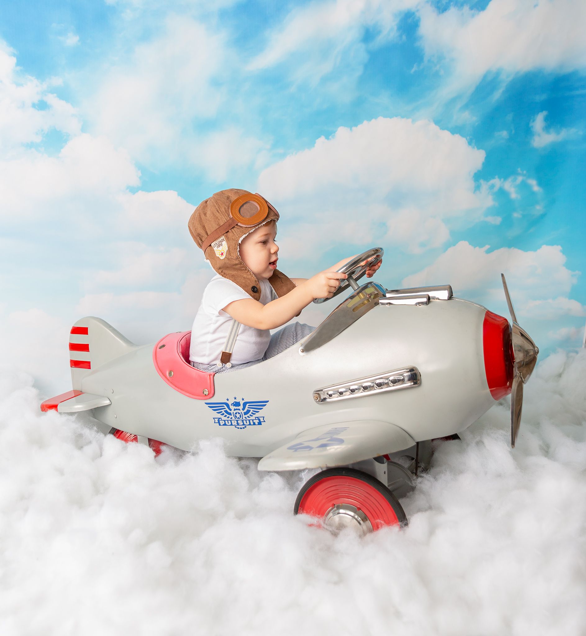 Baby in vintage aviator outfit flying through cloudy blue sky background.