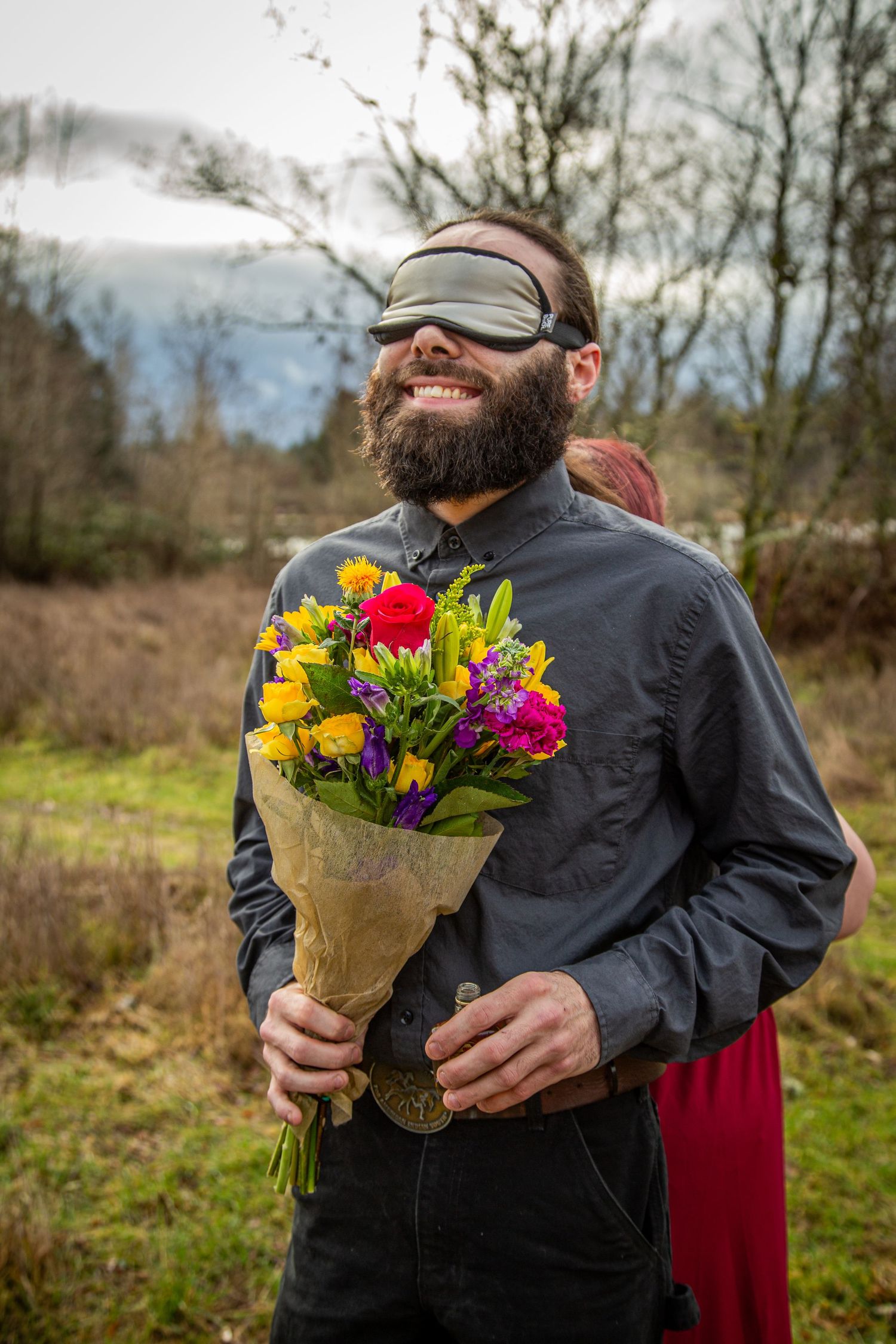 A joyful person wearing a dark jacket holds a colorful bouquet of flowers while standing in a field on an overcast day.