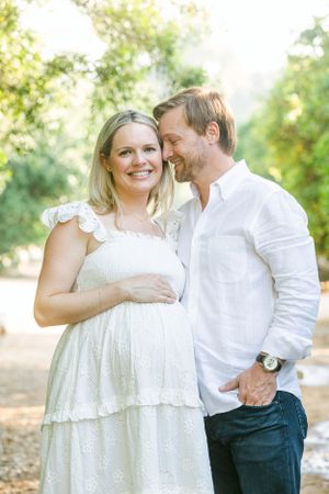 Blond pregnant woman wearing cream dress holding belly while her husband nuzzles her in a garden