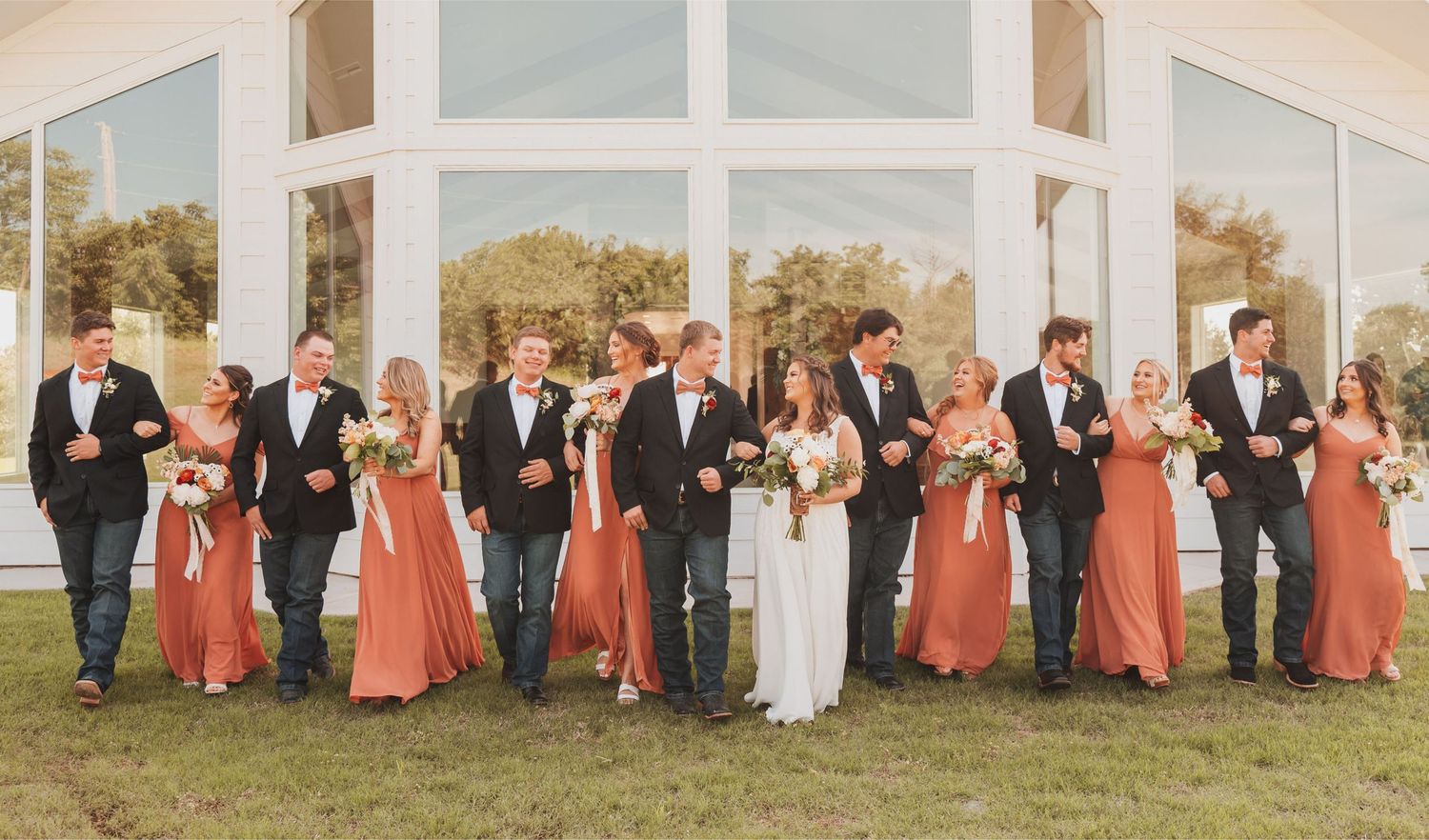 Bride, groom, and their wedding party pictured in front of large windows reflecting blue skies and trees.