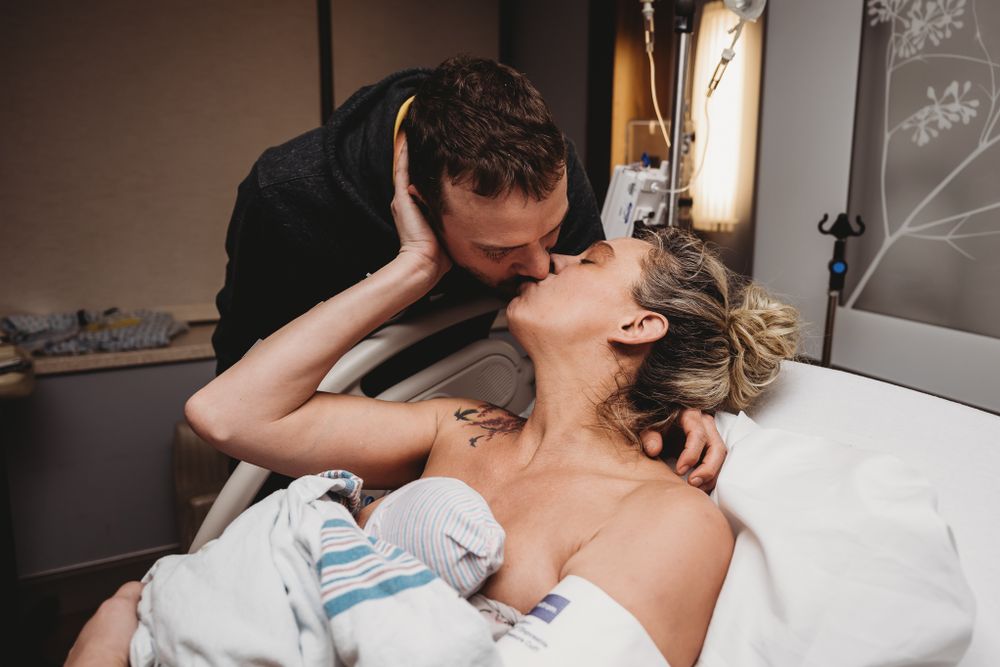 Emotional partner kisses new mother holding swaddled newborn in hospital bed after childbirth.