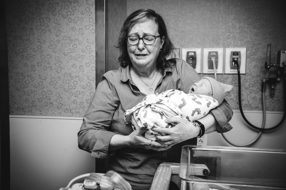 Emotional grandmother cries while holding swaddled newborn baby in hospital room.