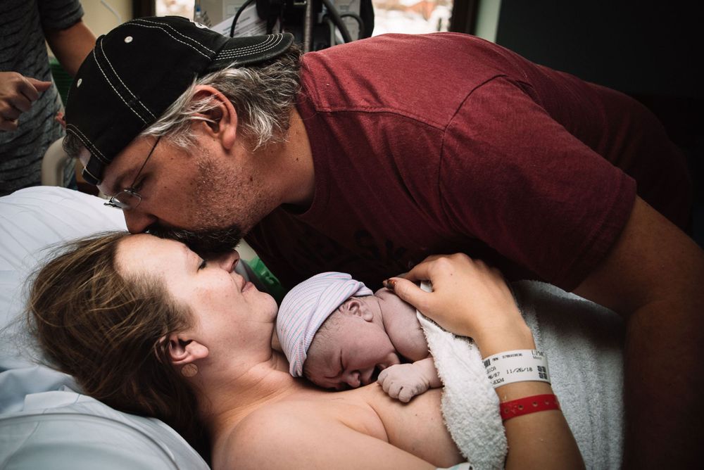Parents share a tender kiss over their newborn baby in a hospital room after birth.
