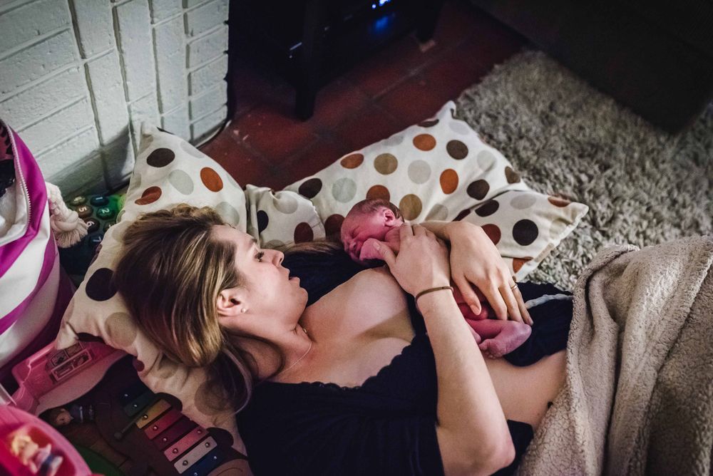 New mother lying on floor cradling newborn baby against her chest at home.