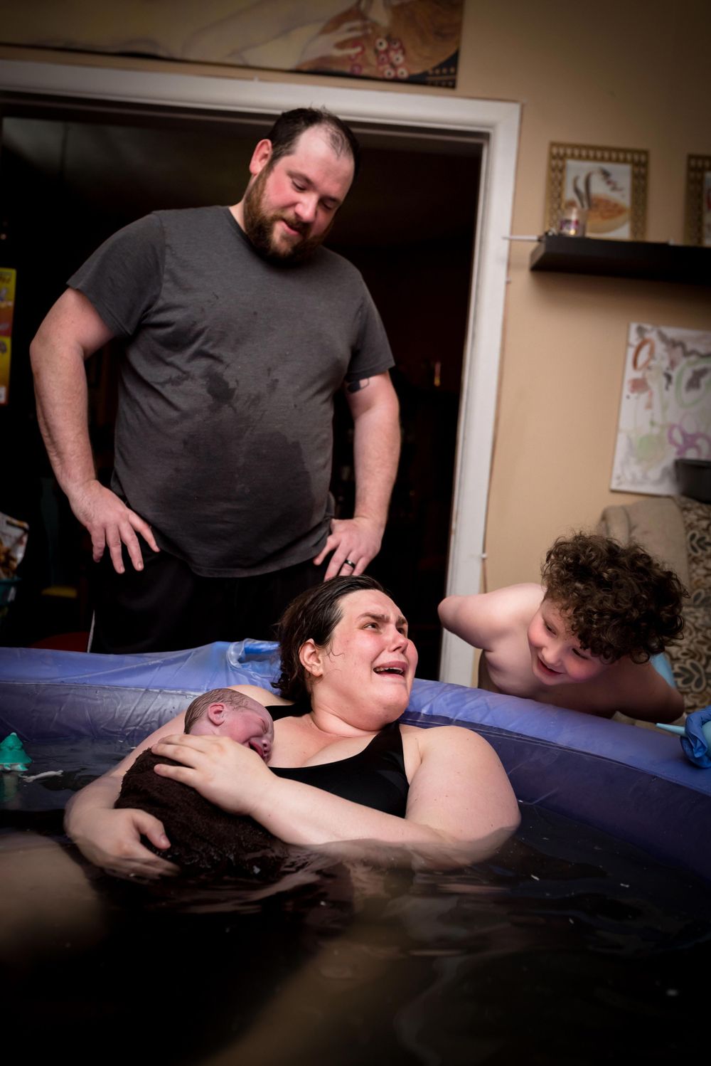 Family witnessing water birth at home, mother holding newborn in inflatable birthing pool.