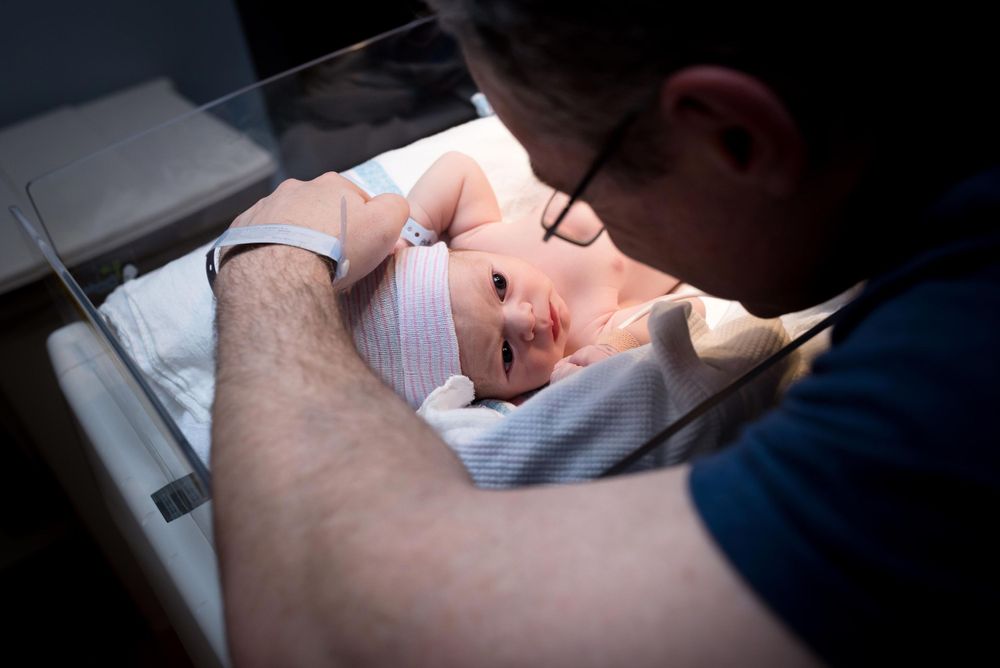 Father gazing lovingly at swaddled newborn baby in hospital bassinet after birth.