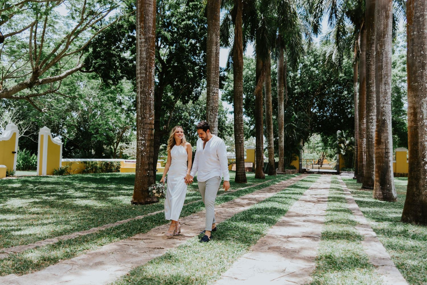 A couple in white outfits walks along a palm tree-lined pathway on a sunny day.