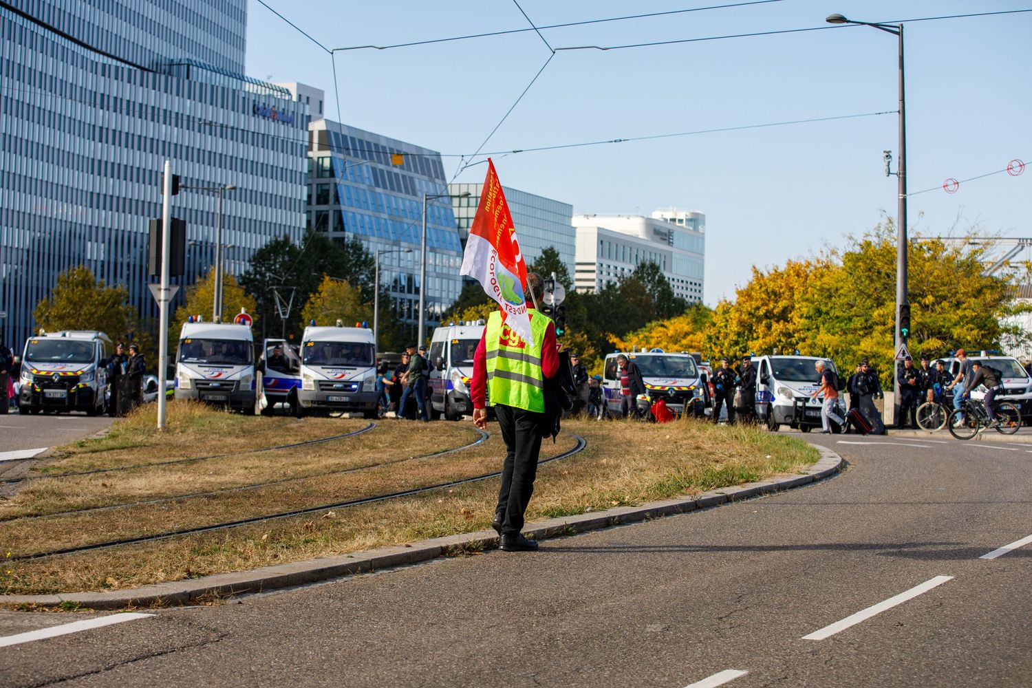 A person in a safety vest carrying a flag walks along a street lined with cars and modern office buildings.