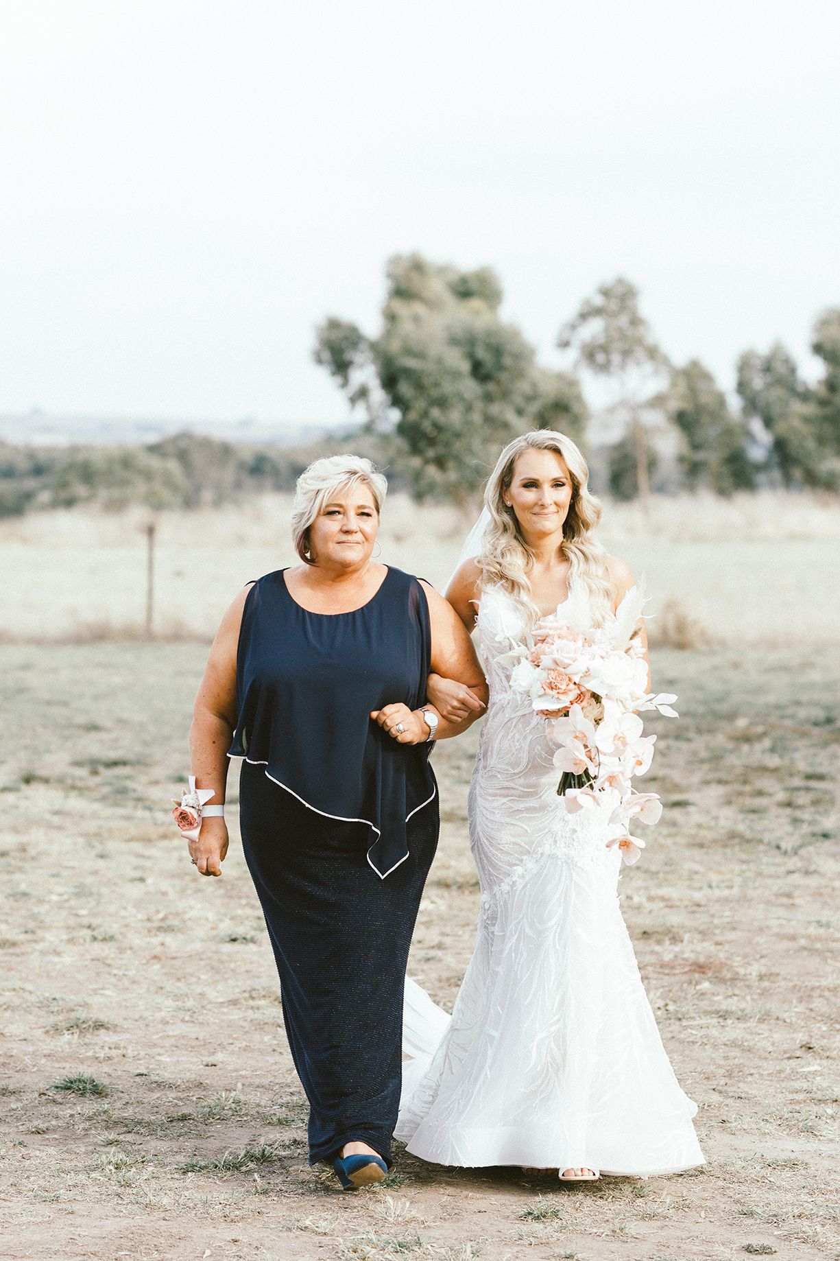 Two women walking arm in arm at an outdoor wedding, one wearing a navy dress and one in a white lace wedding gown.