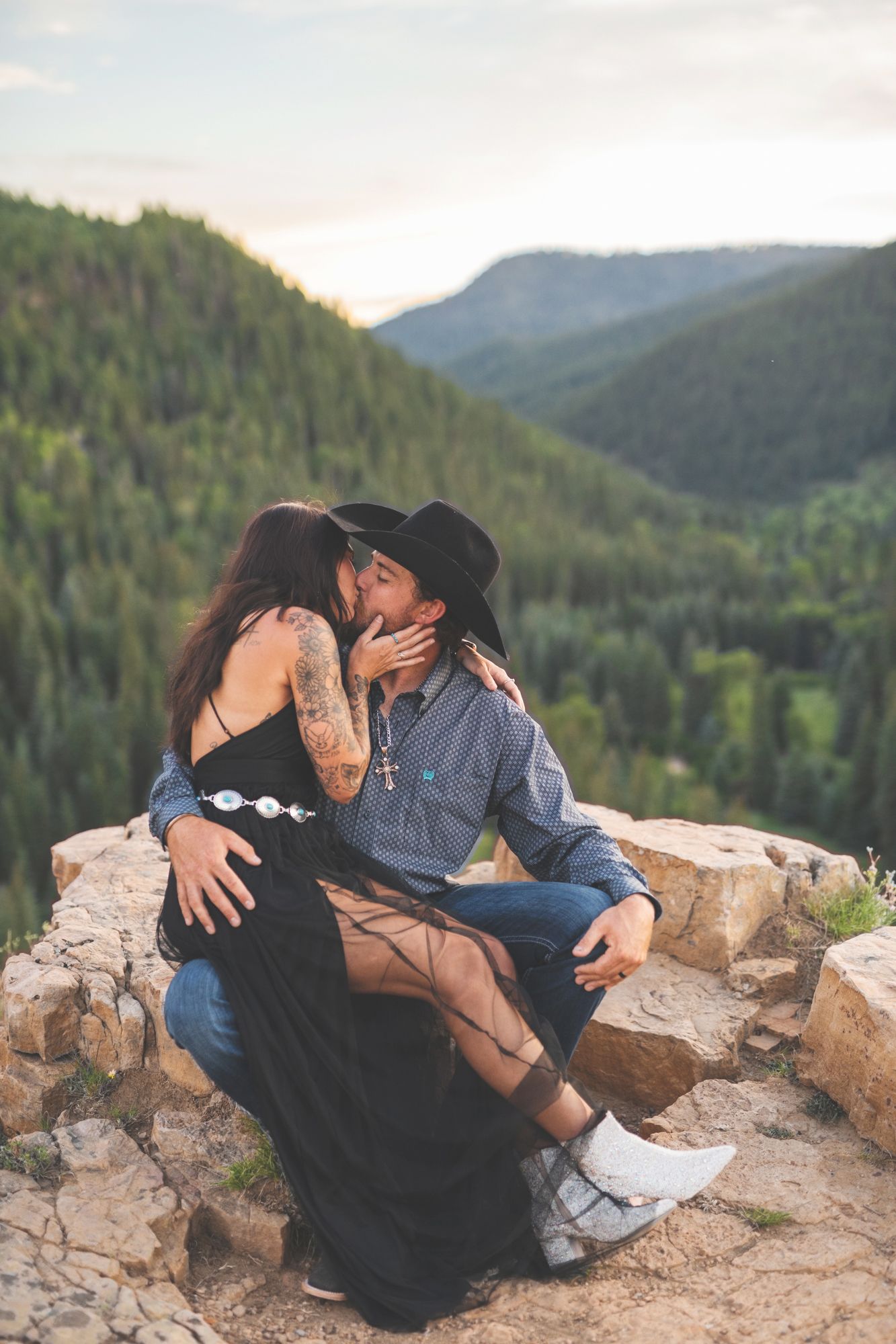 woman in black dress and man in cowboy hat kissing on a cliffside