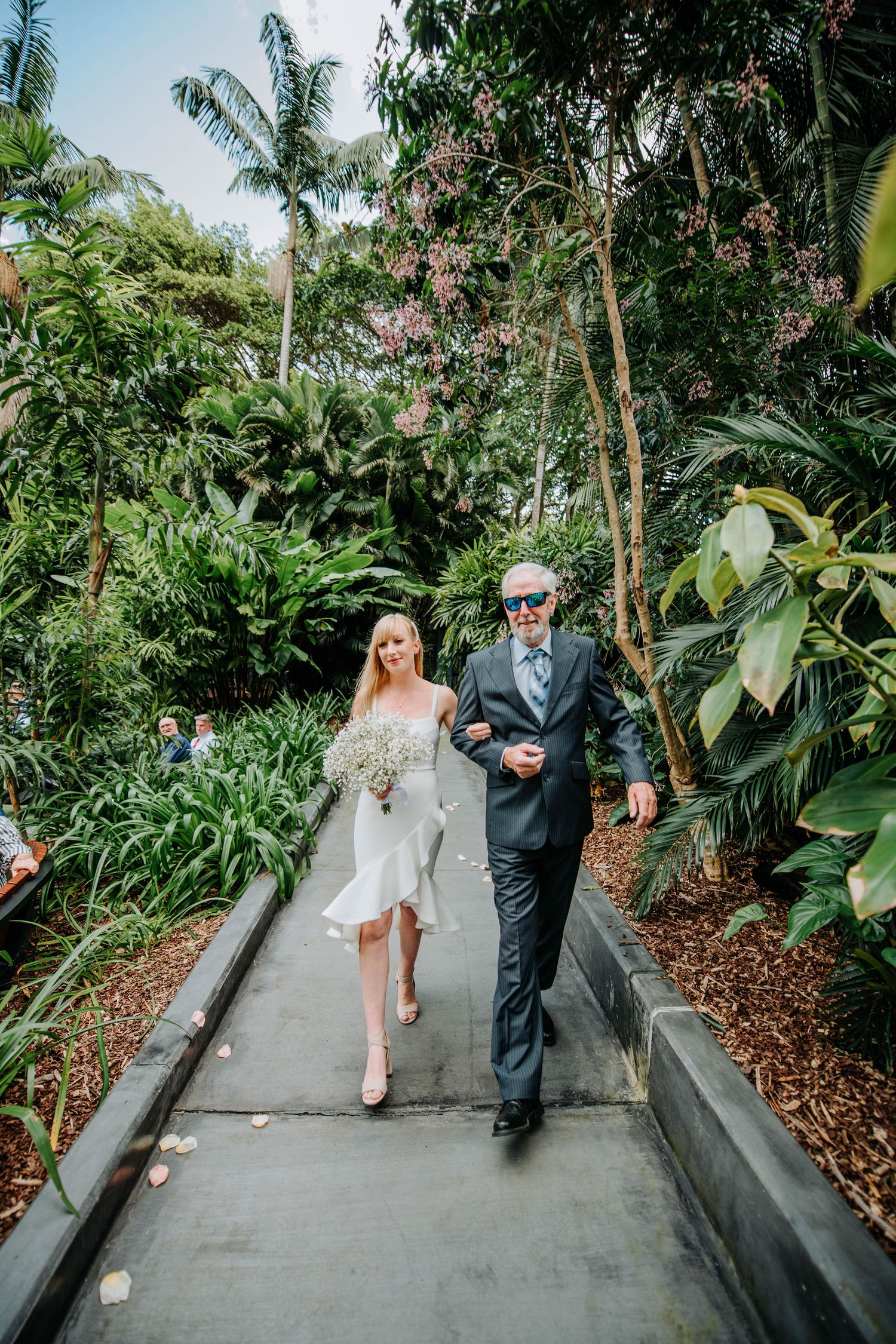 Couple walking along concrete path surrounded by lush tropical greenery and palm trees.