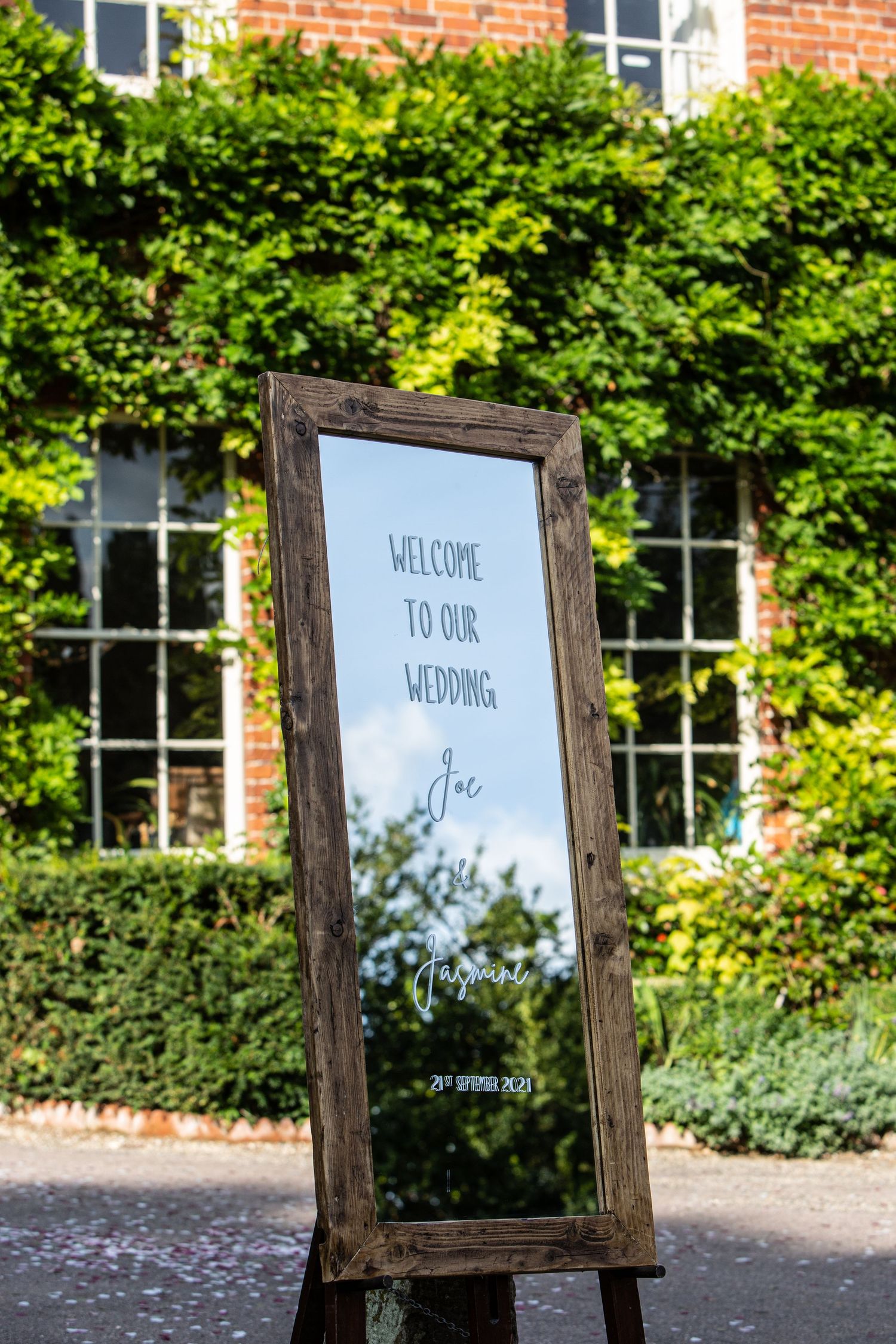 Rustic wooden welcome sign with greenery backdrop and brick building in background.