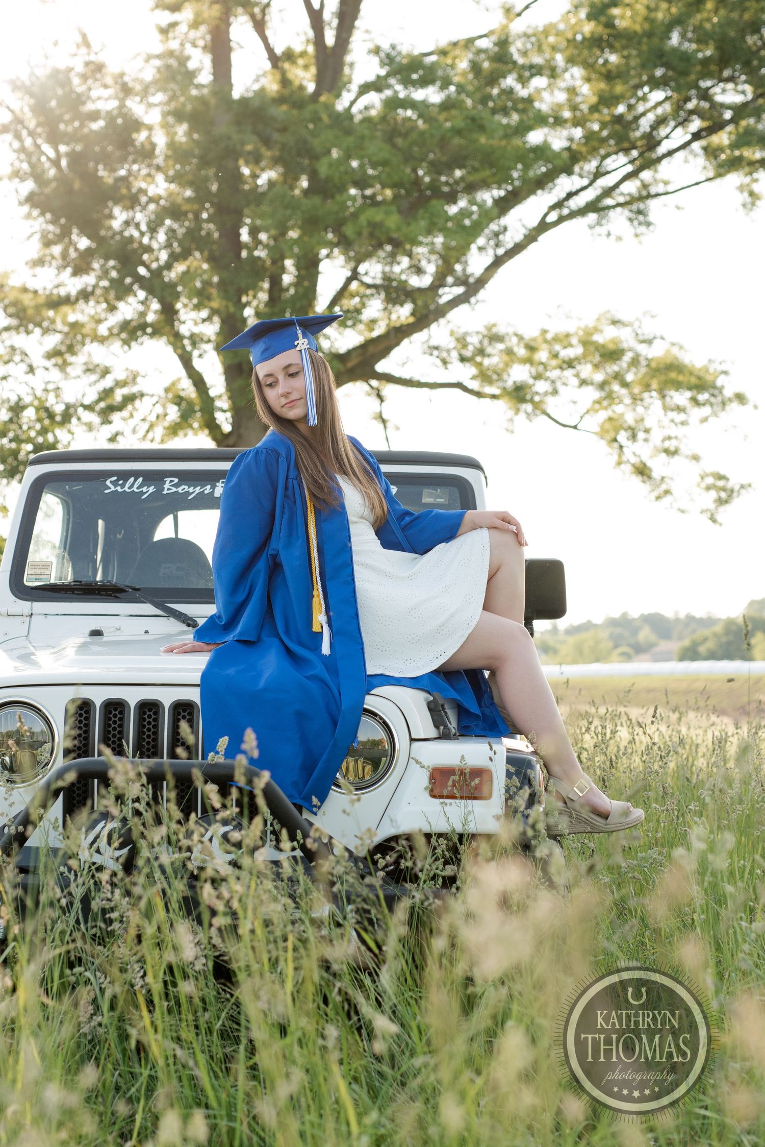 Graduation photo featuring blue cap and gown against white Jeep in golden sunlit meadow.