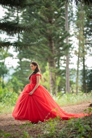 Elegant young woman in a flowing red off-shoulder ballgown standing gracefully among tall pine trees outdoors.