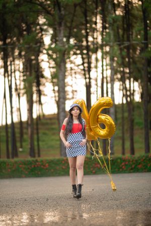 Teen girl holding gold number 16 balloons walking through a pine forest in a checkered skirt and red top.